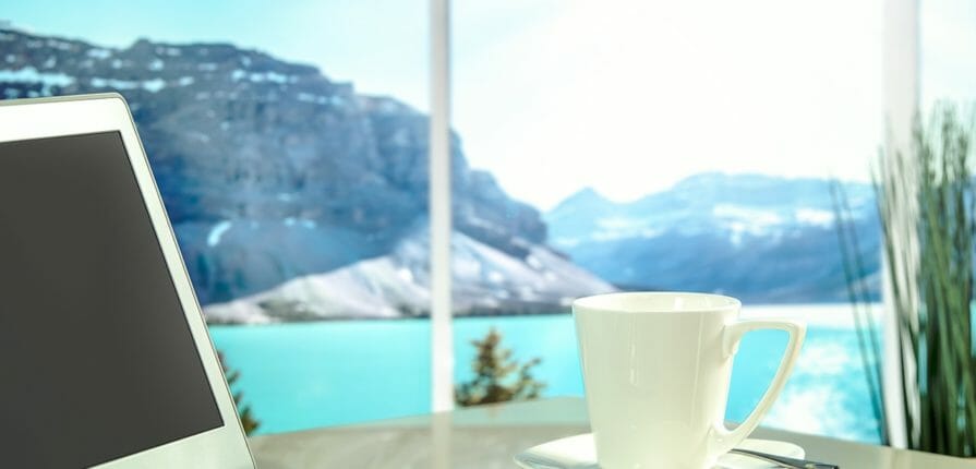 Cup of coffee and laptop in a sunroom equipped with windows outfitted with Climate Seal thermal window inserts overlooks a lake at the base of a mountain range.