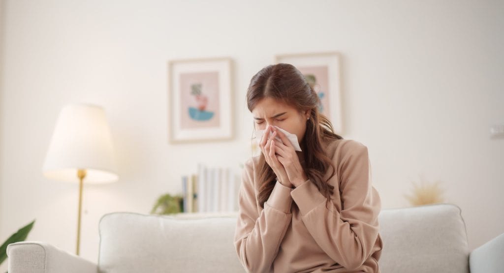 Young Asian woman sits on a couch, sneezing and holding a tissue to her nose while an air purifier operates nearby to improve air quality.